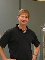 Man in black polo smiling standing in front of an instrumentation for research